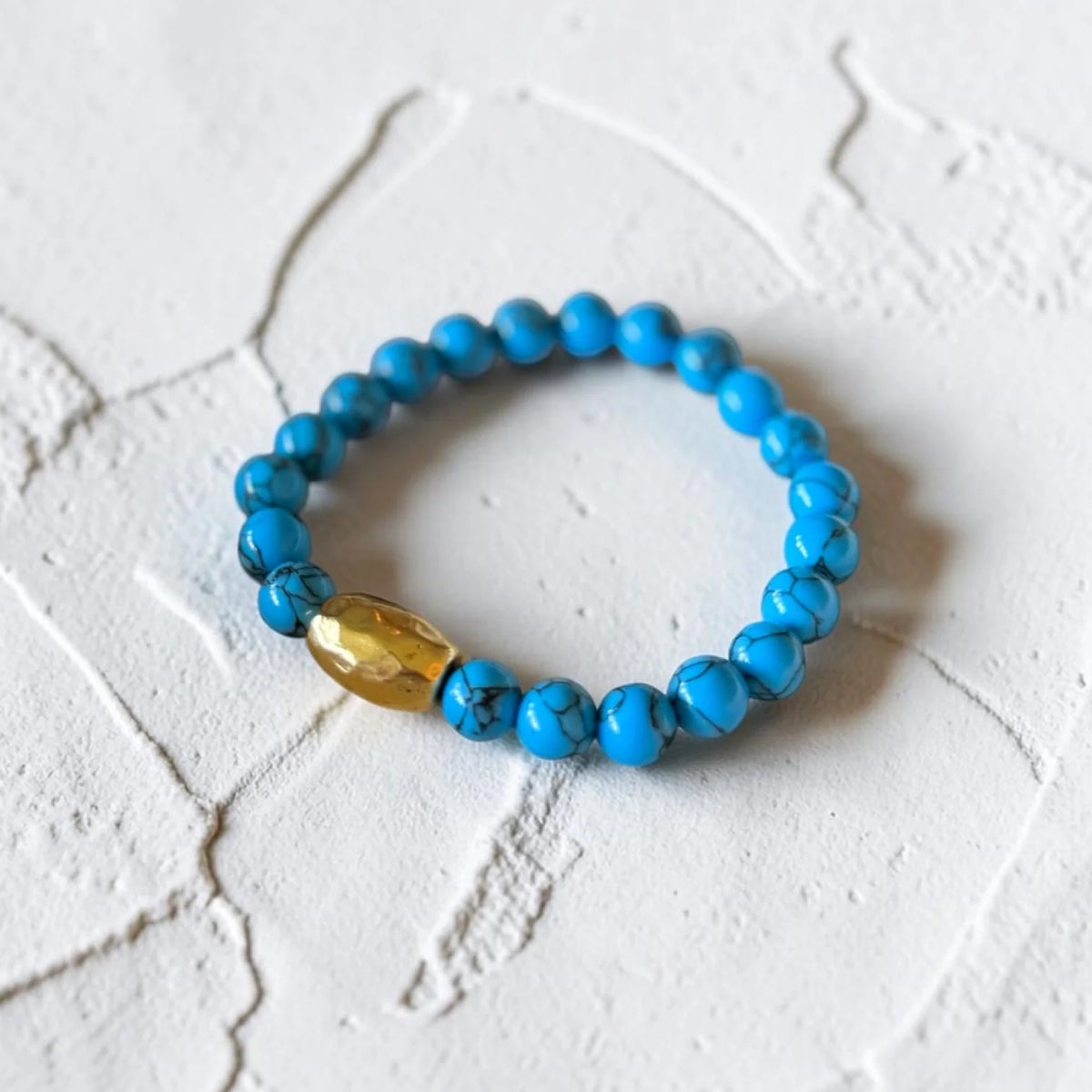 A beaded stretch bracelet featuring round turquoise-blue marbled beads and a single faceted gold accent bead, displayed on a textured white surface.