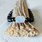 A beaded bracelet with glossy black beads and a central iridescent white bead, displayed on a textured beige woven stand against a light neutral background.