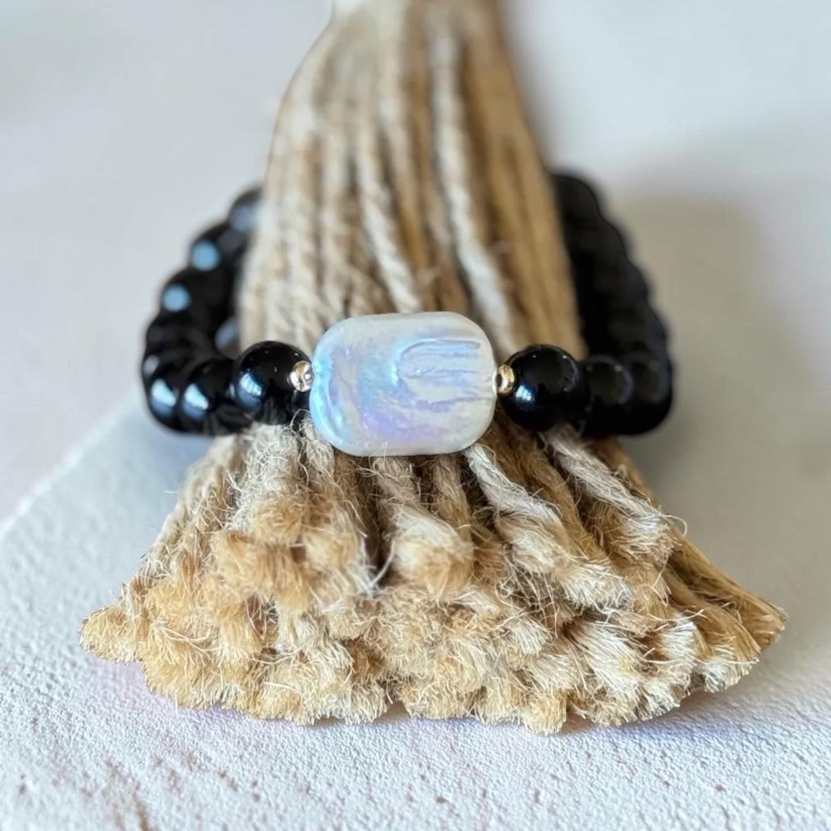 A beaded bracelet with glossy black beads and a central iridescent white bead, displayed on a textured beige woven stand against a light neutral background.