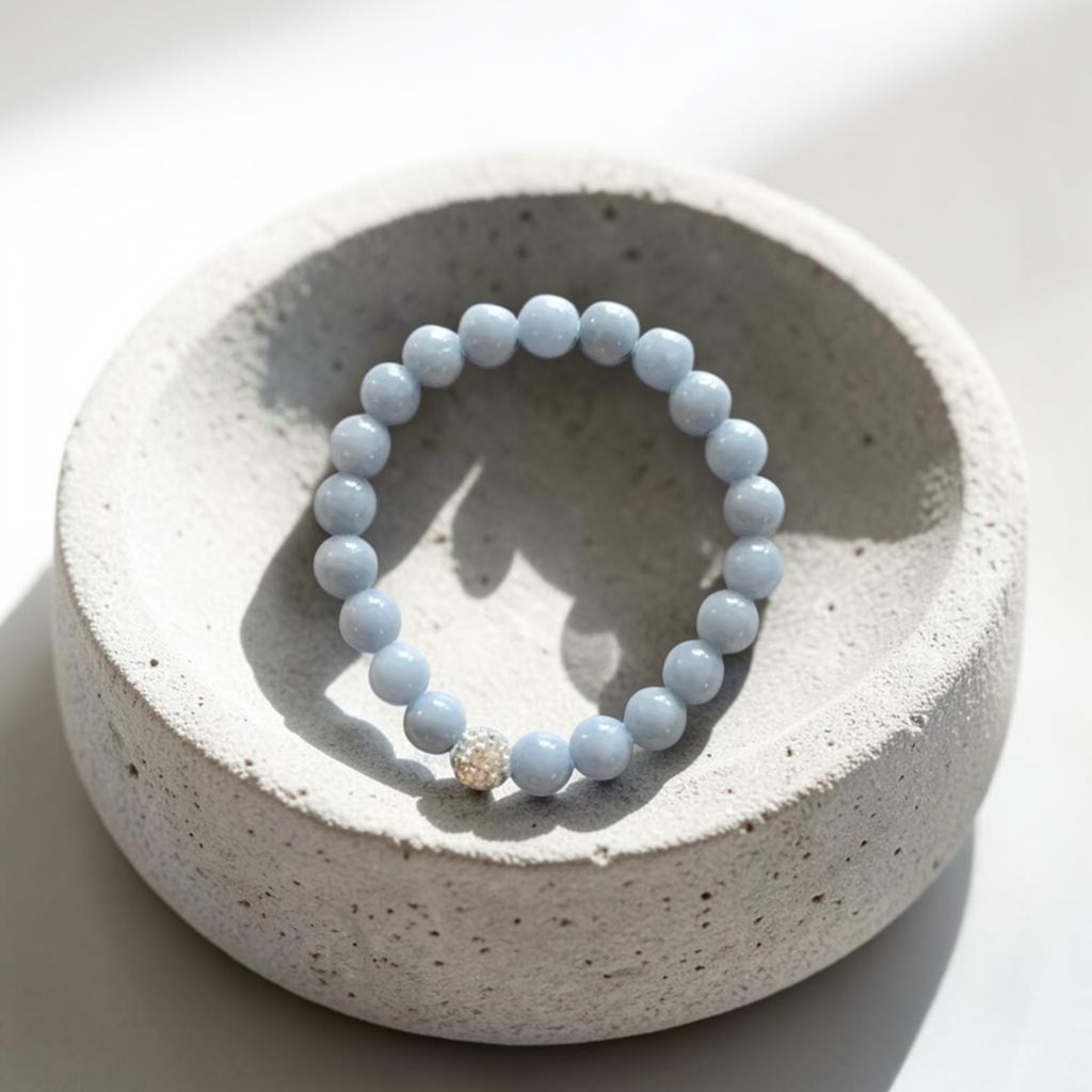 A light blue beaded bracelet with a small gold accent bead, displayed on a textured light gray ceramic dish against a bright white background.