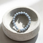 A light blue beaded bracelet with a small gold accent bead, displayed on a textured light gray ceramic dish against a bright white background.