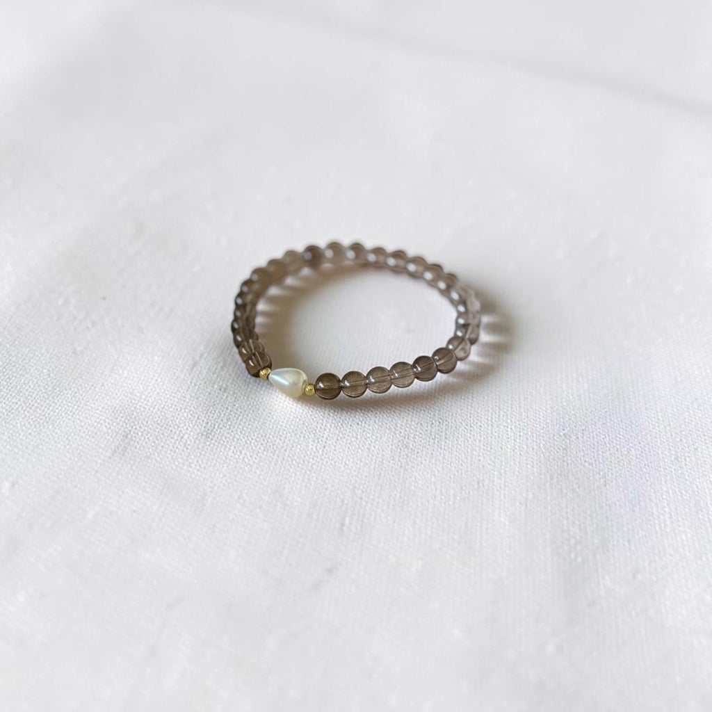 A delicate beaded bracelet with see through brown beads and a tiny iridescent accent, resting on a soft white fabric background.