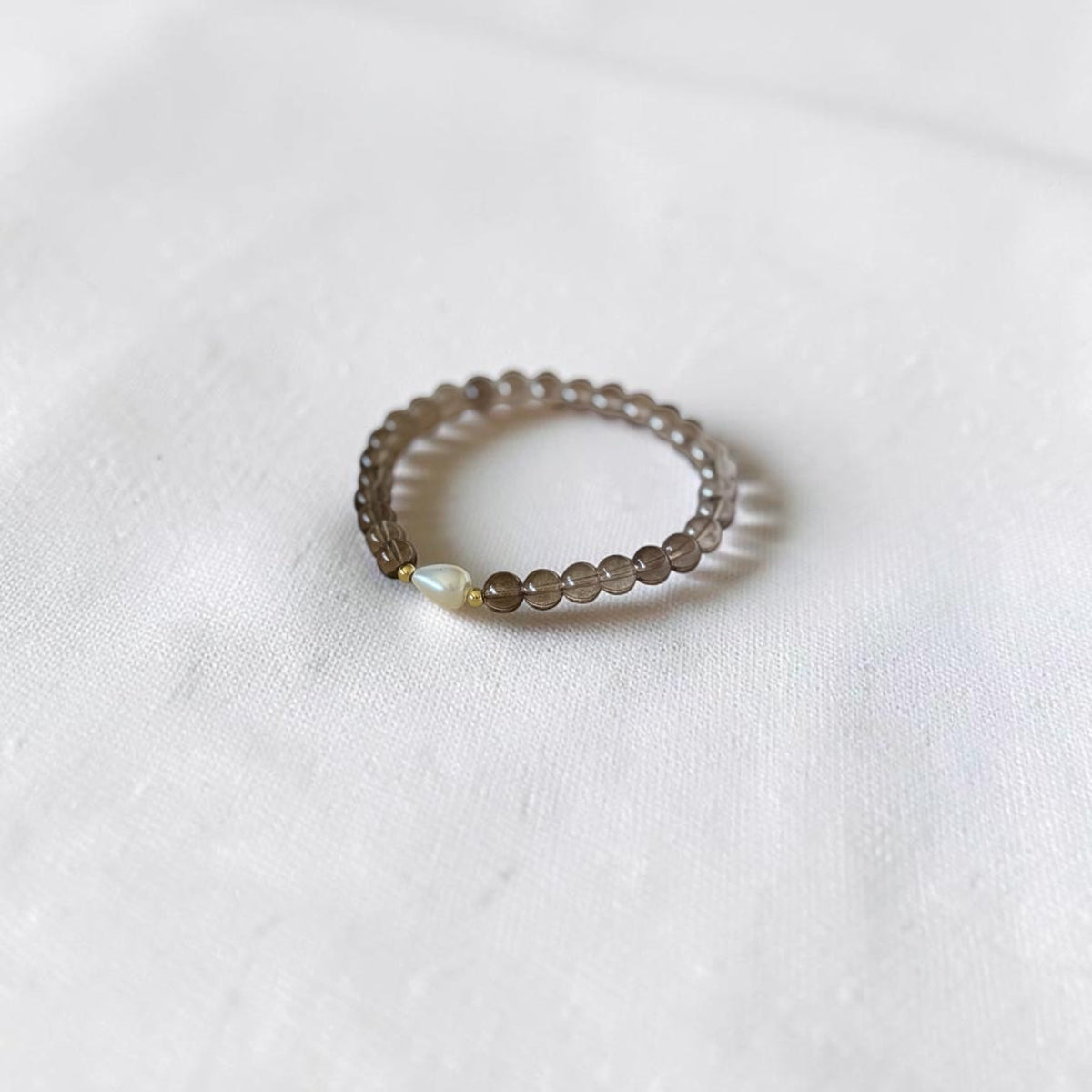 A delicate beaded bracelet with see through brown beads and a tiny iridescent accent, resting on a soft white fabric background.