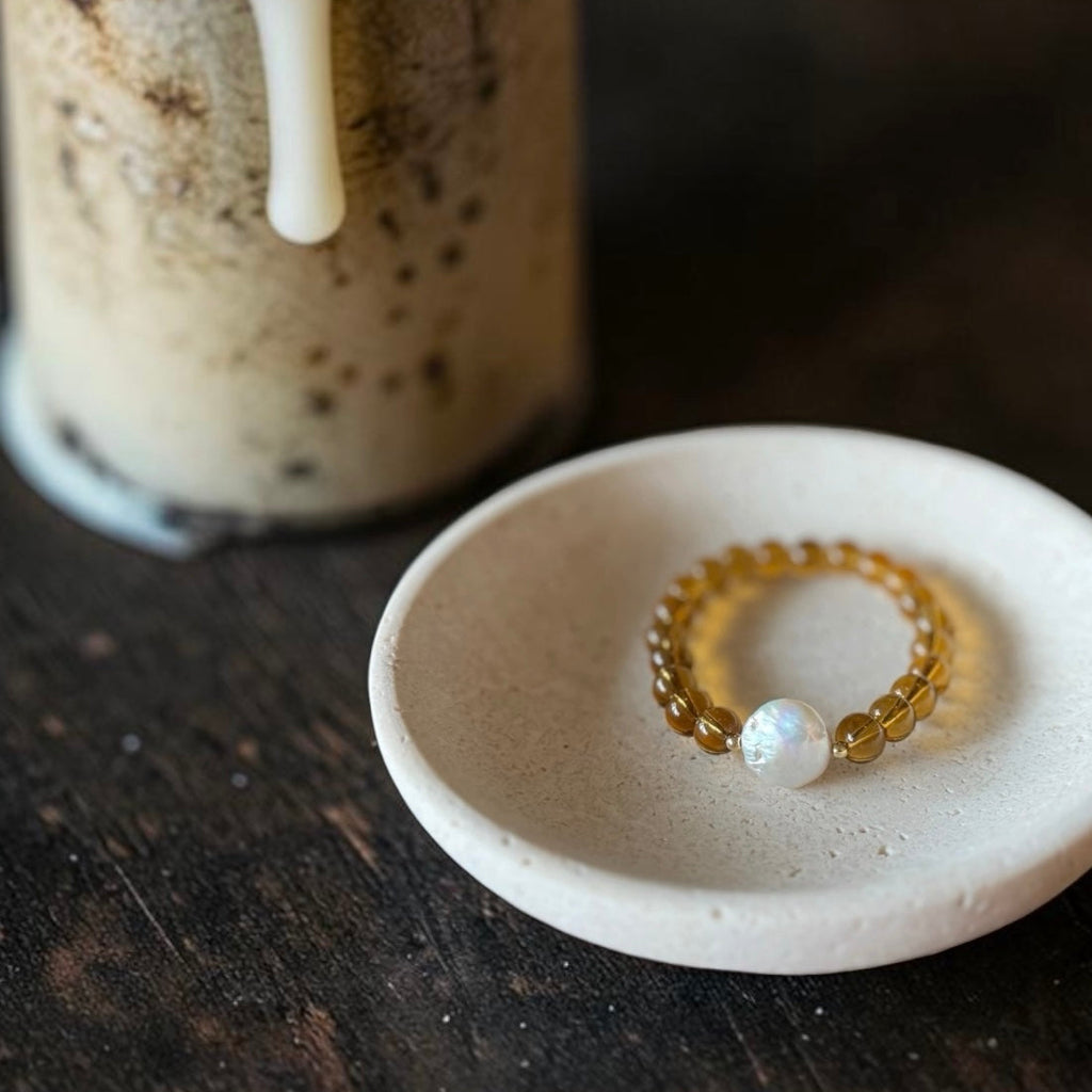 A beaded bracelet with amber-yellow translucent beads and a white pearl-like center bead, displayed on a small round ceramic dish over a dark wooden surface, with a blurred drink in the background.