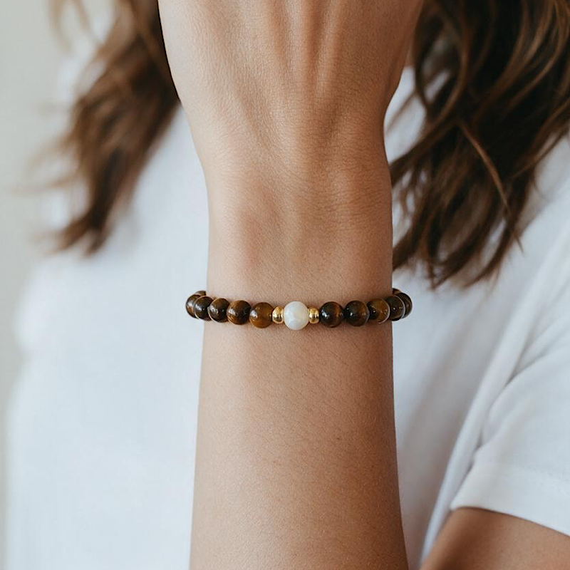 A close-up of a person's wrist wearing a beaded bracelet made of brown and gold beads with a single white bead accent, against a soft white shirt background.