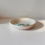 A beaded bracelet with light on green beads and a small white pearl accent, displayed on a textured cream ceramic dish against a warm, minimalist background.