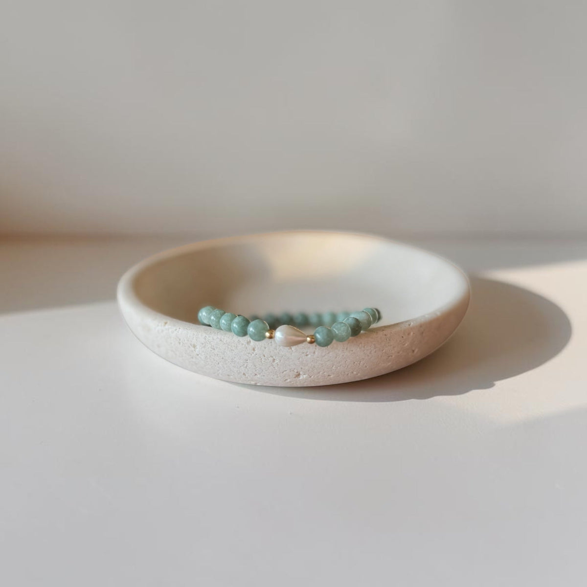 A beaded bracelet with light on green beads and a small white pearl accent, displayed on a textured cream ceramic dish against a warm, minimalist background.