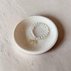 A beaded bracelet with a small gold bow charm, displayed inside a small speckled ceramic dish on a textured beige surface.