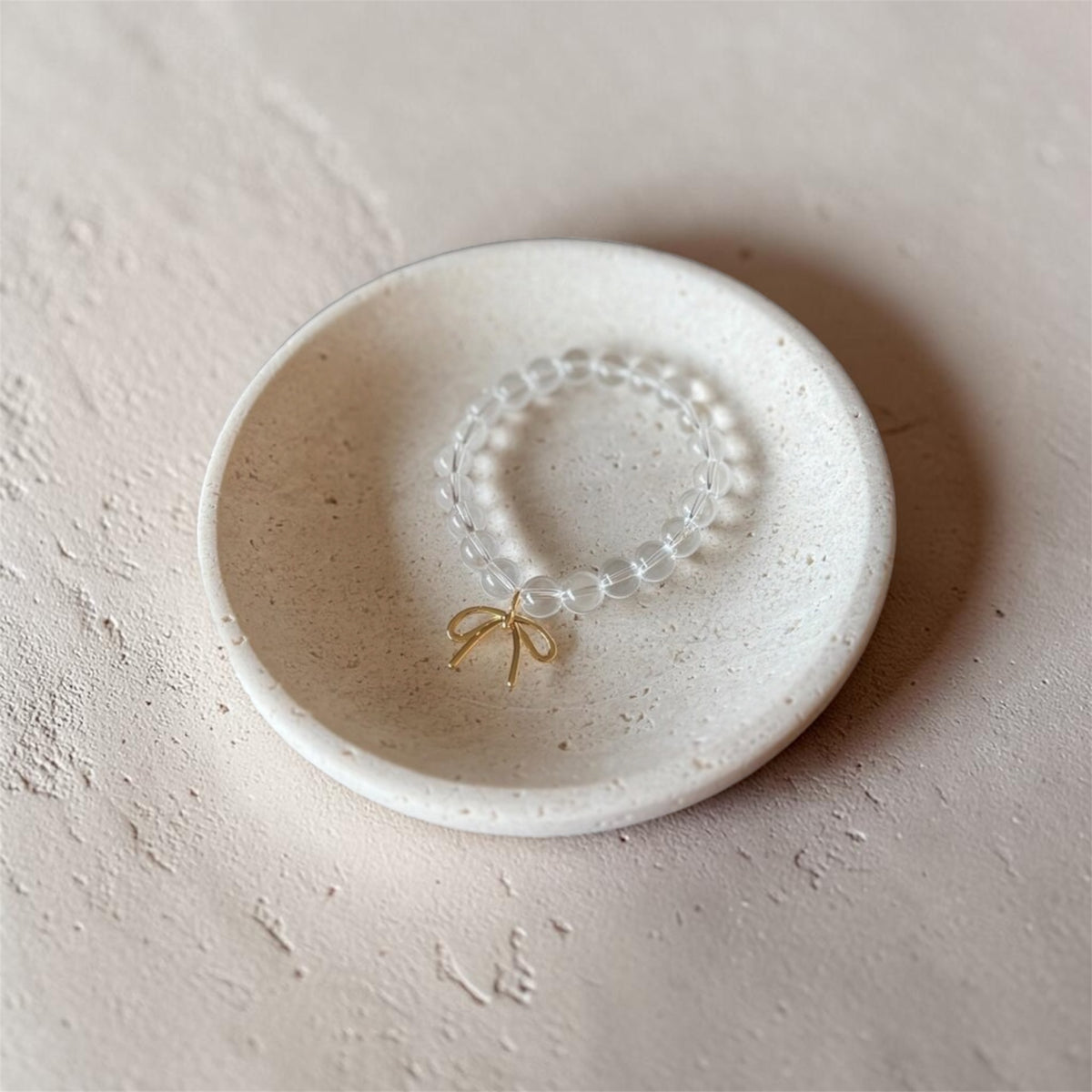 A beaded bracelet with a small gold bow charm, displayed inside a small speckled ceramic dish on a textured beige surface.