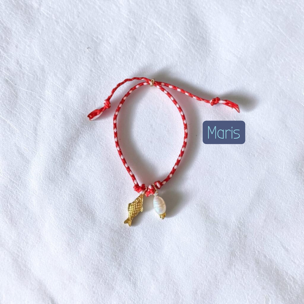 A red and white patterned string bracelet with a small gold fish charm and a white shell charm, displayed on a textured white fabric background with a blue label reading 'Maris'.
