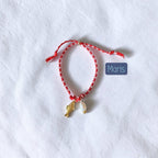 A red and white patterned string bracelet with a small gold fish charm and a white shell charm, displayed on a textured white fabric background with a blue label reading 'Maris'.