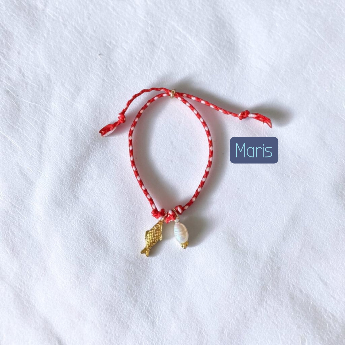 A red and white patterned string bracelet with a small gold fish charm and a white shell charm, displayed on a textured white fabric background with a blue label reading 'Maris'.