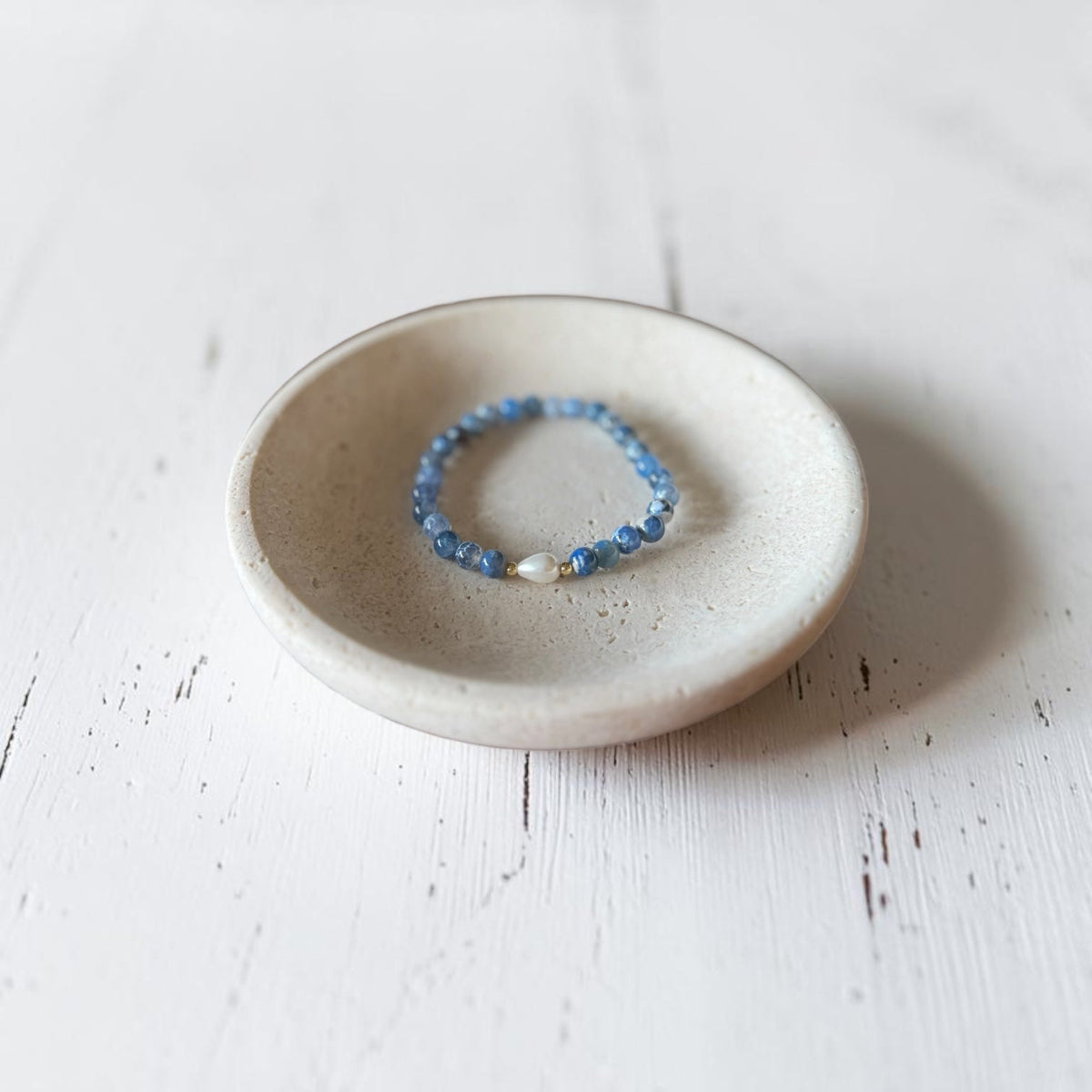 A beaded stretch bracelet with blue beads and a small white pearl accent, displayed inside a small round ceramic dish on a white wooden surface.