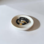 A pair of beaded bracelets, one in matte black and one with black, gold, and blue patterned beads, displayed in a small round ceramic dish on a white surface.