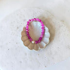 A beaded stretch bracelet made of bright pink faceted beads with a small white pearl accent, displayed on a small scalloped dish over a textured white surface.
