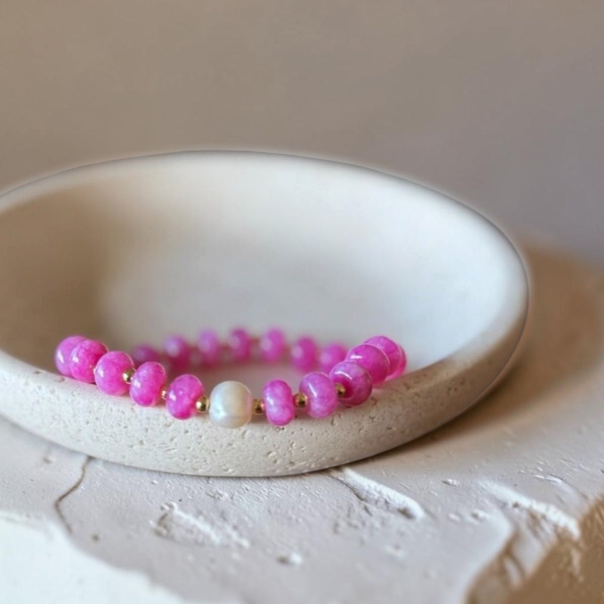 A beaded stretch bracelet with bright pink round beads, small gold spacer beads, and one white pearl-like bead, displayed inside a shallow off-white ceramic dish.