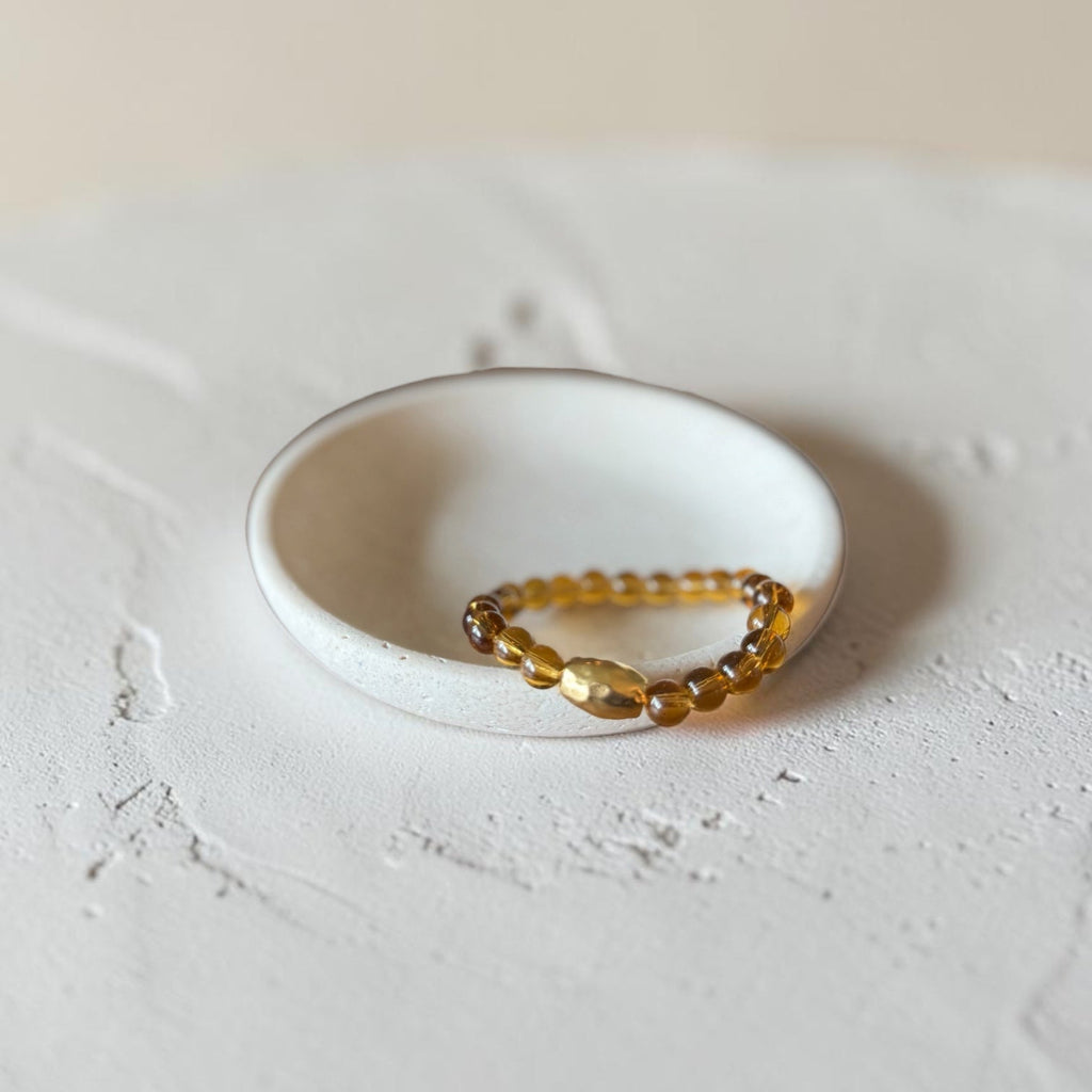 A beaded stretch bracelet with amber and gold-toned beads, displayed in a small white ceramic dish on a textured light gray surface.