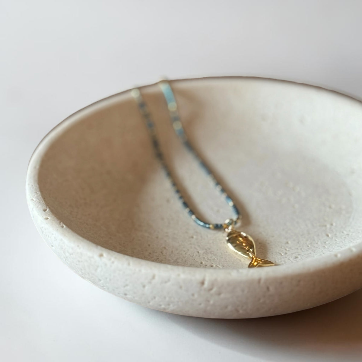 A delicate necklace with a small gold fish pendant and light blue beads, resting inside a textured beige ceramic dish on a white surface.