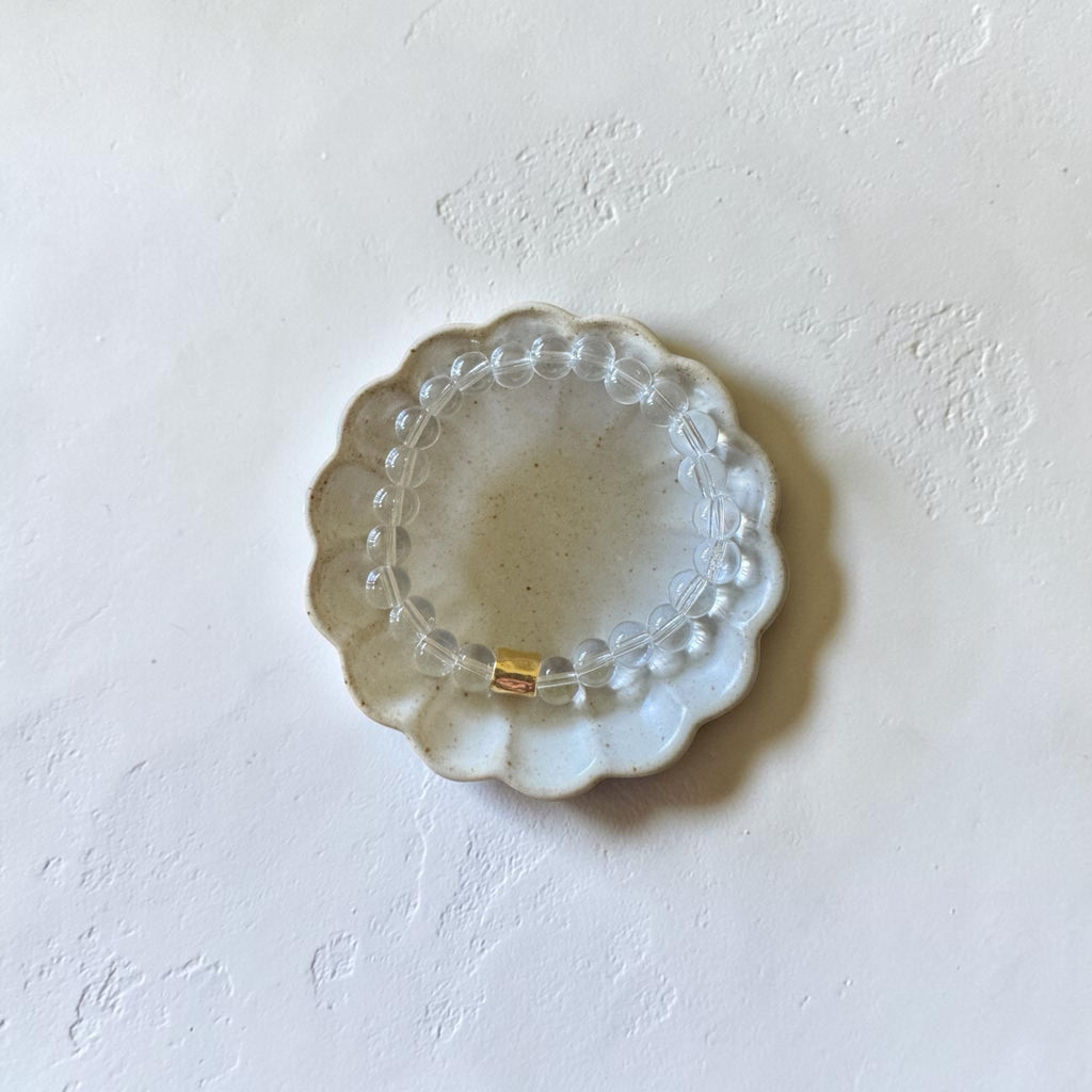 A clear beaded bracelet with a small gold accent ring, displayed on a scalloped clear glass dish against a textured off-white background.