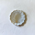 A clear beaded bracelet with a small gold accent ring, displayed on a scalloped clear glass dish against a textured off-white background.
