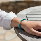 A turquoise beaded bracelet with a gold-toned central accent worn on a person's wrist, resting on a gray wooden table.