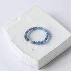 A beaded stretch bracelet with blue and gray beads and a small white pearl accent, displayed on a textured white stone block against a light gray background.