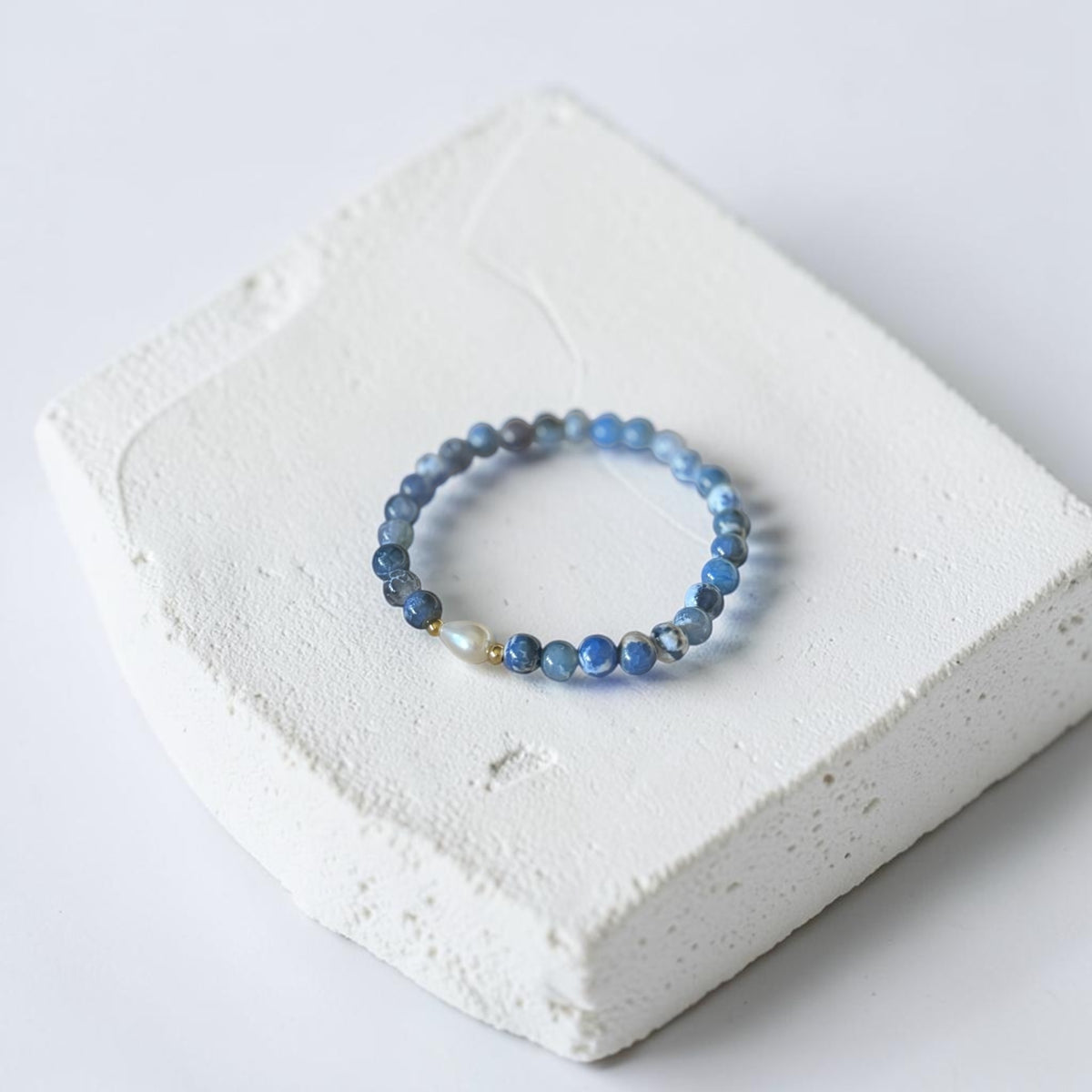 A beaded stretch bracelet with blue and gray beads and a small white pearl accent, displayed on a textured white stone block against a light gray background.