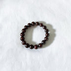 A dark brown beaded bracelet made of polished round beads, arranged in a circle on a textured white fabric background.