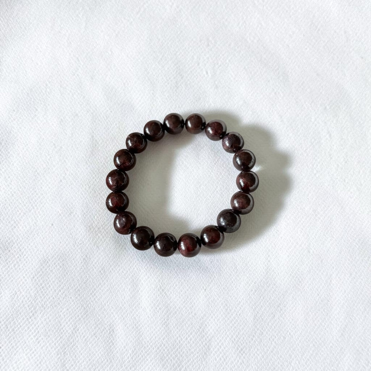 A dark brown beaded bracelet made of polished round beads, arranged in a circle on a textured white fabric background.
