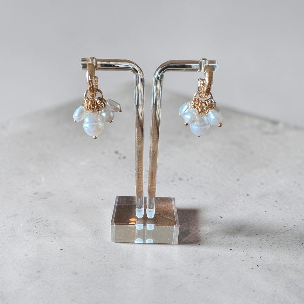 A pair of gold hoop earrings with small white pearl clusters hanging from the hoops, displayed on a clear acrylic jewelry stand against a light gray background.