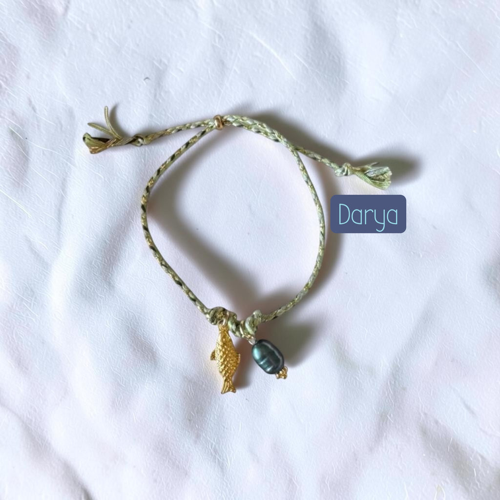 A handmade braided string bracelet in a light olive green color, featuring a small gold fish charm and a dark gray teardrop bead. The bracelet is displayed on a wrinkled white fabric background with a blue label overlay reading 'Darya'.