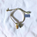 A handmade braided string bracelet in a light olive green color, featuring a small gold fish charm and a dark gray teardrop bead. The bracelet is displayed on a wrinkled white fabric background with a blue label overlay reading 'Darya'.