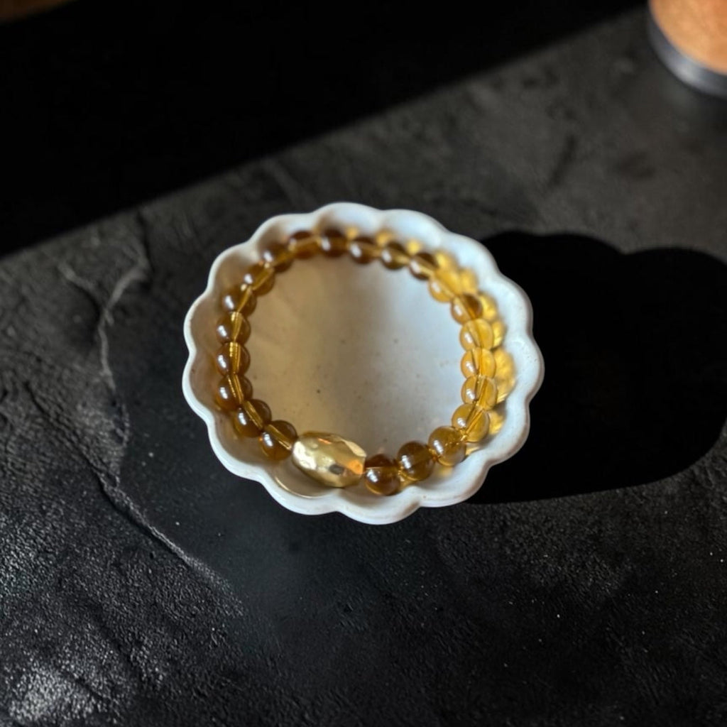 A beaded bracelet made of translucent amber-yellow glass beads with a larger polished oval bead, displayed inside a small white scalloped dish on a dark textured surface.