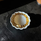 A beaded bracelet made of translucent amber-yellow glass beads with a larger polished oval bead, displayed inside a small white scalloped dish on a dark textured surface.