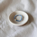A beaded stretch bracelet with blue and white beads resting in a small, round, textured ceramic dish on a soft, draped white fabric background.