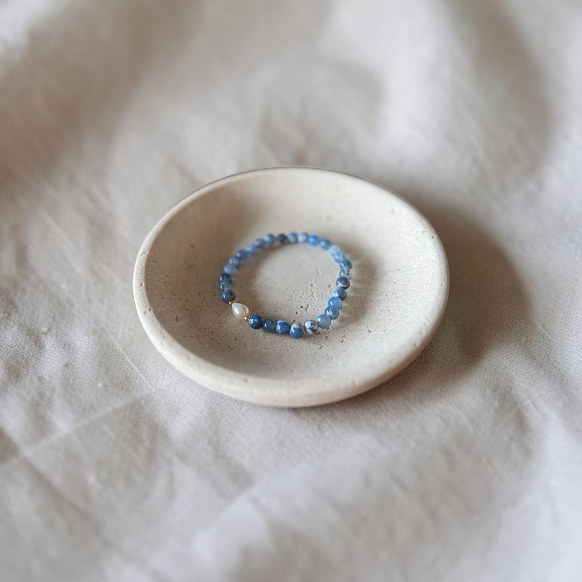 A beaded stretch bracelet with blue and white beads resting in a small, round, textured ceramic dish on a soft, draped white fabric background.