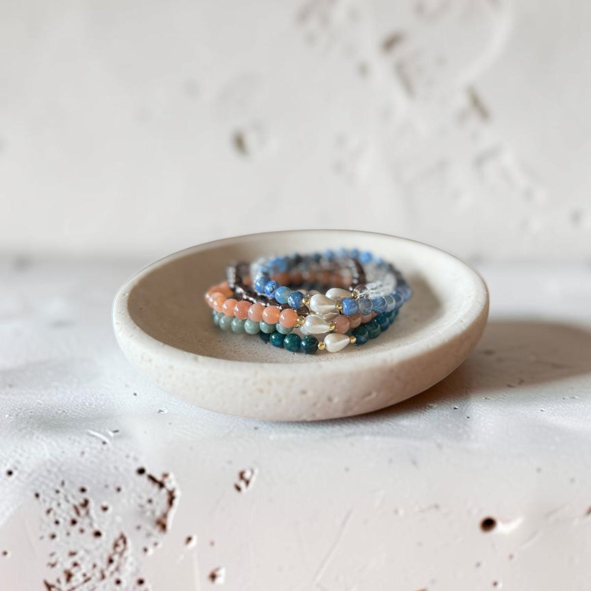 A stack of beaded bracelets in blue, peach, and white, displayed on a small round ceramic dish against a textured off-white background.