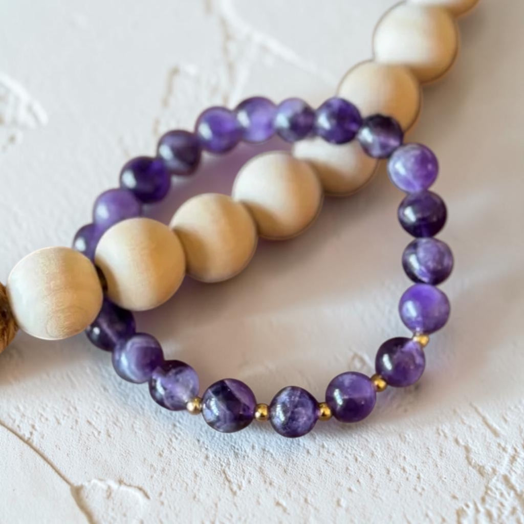 Two beaded bracelets displayed on a textured white surface. One bracelet features round purple amethyst beads separated by small gold spacer beads, while the other features larger natural light wood beads.