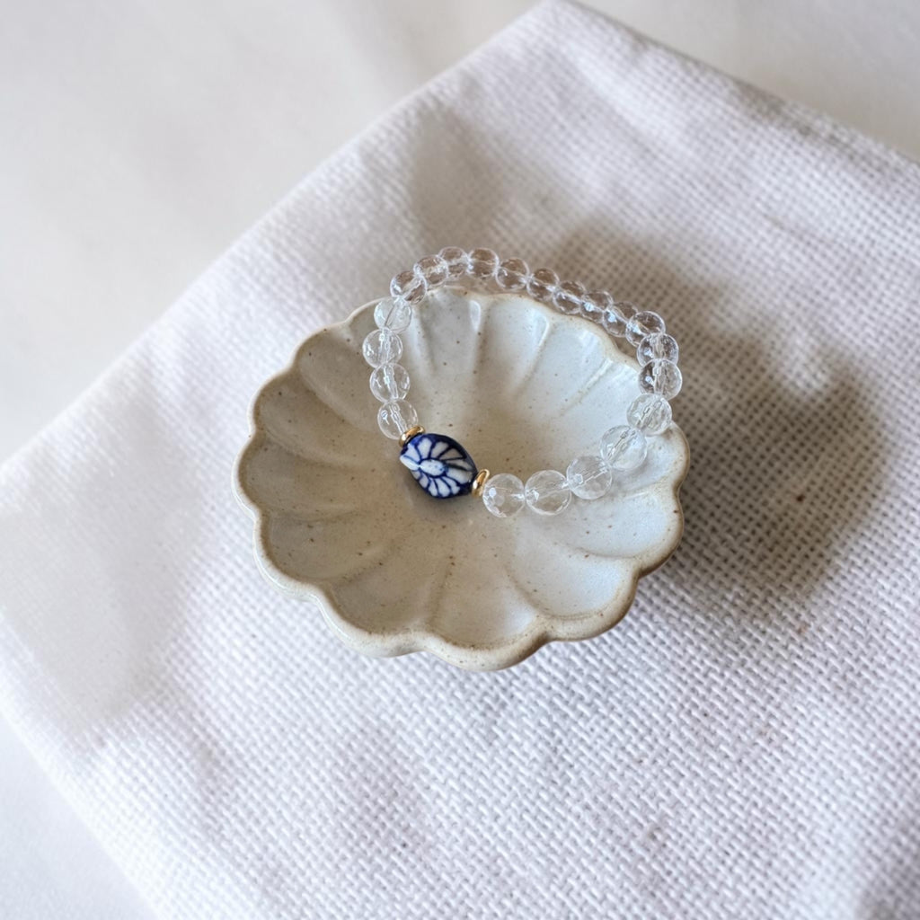 A clear beaded bracelet with a central blue and white floral bead, displayed on a small scalloped ceramic dish over a white textured fabric background.