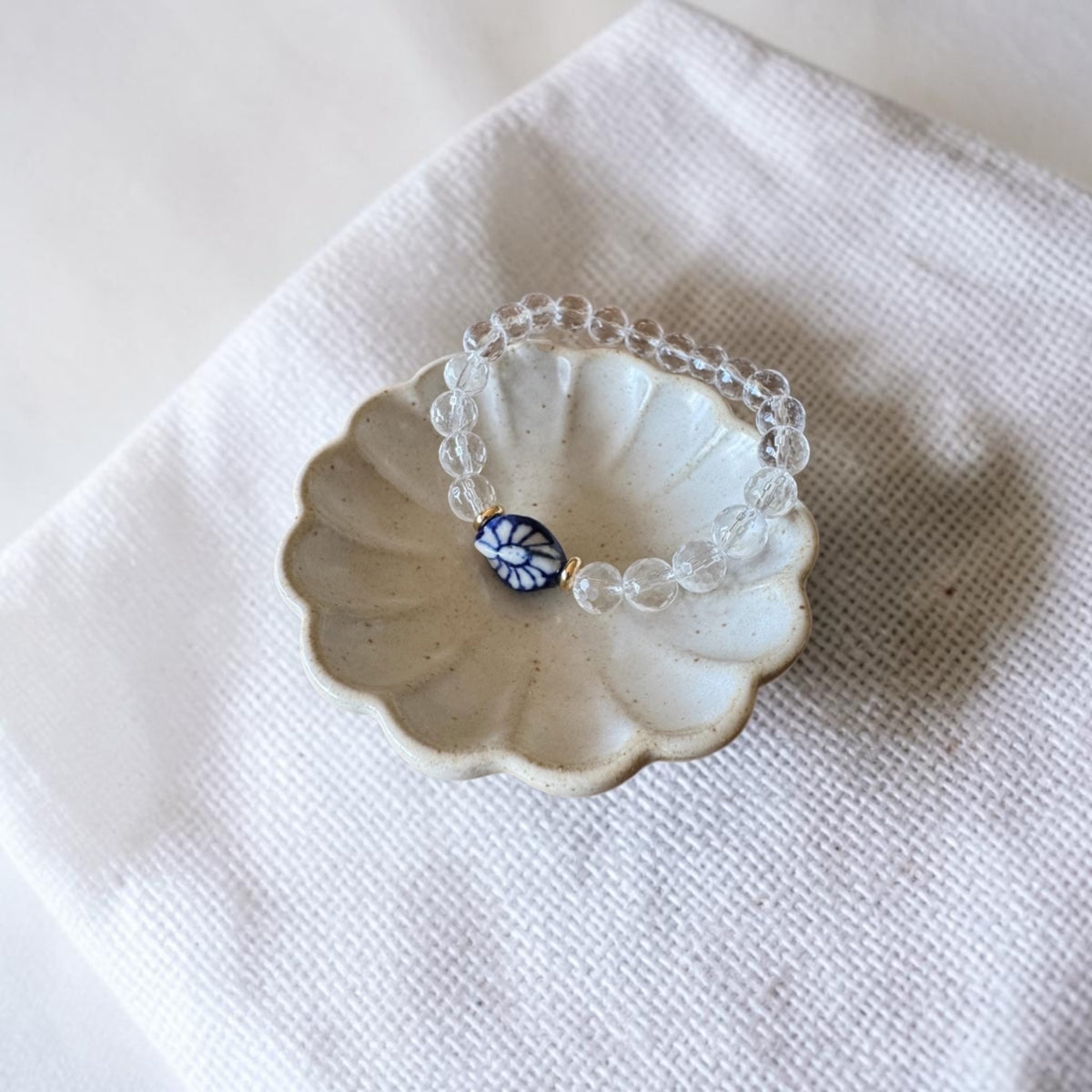 A clear beaded bracelet with a central blue and white floral bead, displayed on a small scalloped ceramic dish over a white textured fabric background.