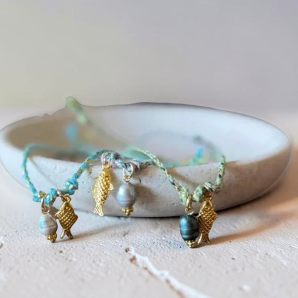 Three delicate braided bracelets with gold-toned shell charms and blue-gray beads, displayed on a small white ceramic dish against a light neutral background.