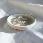 A delicate beaded bracelet with white, black, and metallic beads resting on a small round ceramic dish, set against a draped white fabric background with soft shadows.