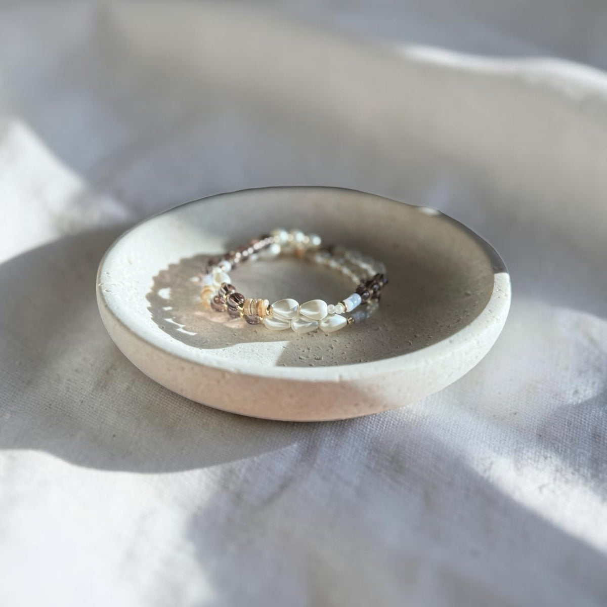 A delicate beaded bracelet with white, black, and metallic beads resting on a small round ceramic dish, set against a draped white fabric background with soft shadows.