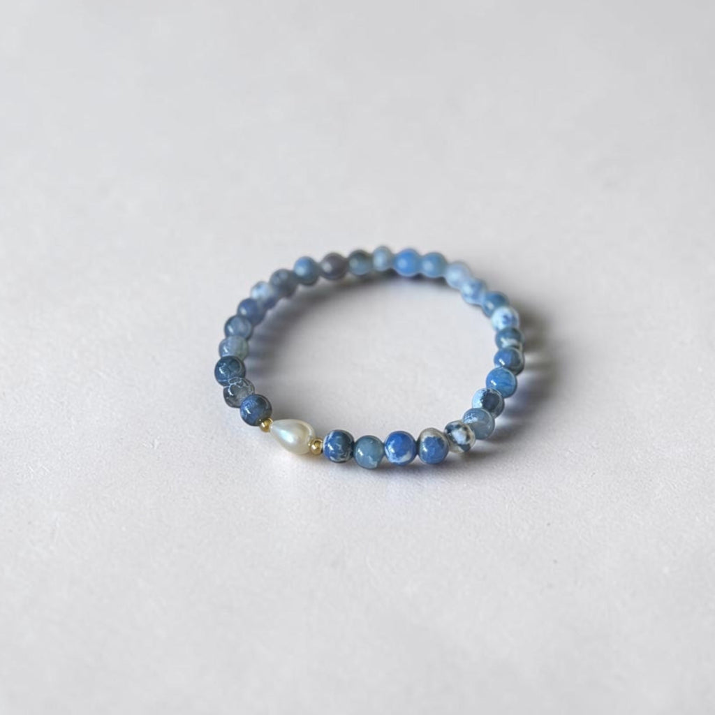 A beaded stretch bracelet featuring small blue and gray beads with a single white teardrop pearl accent, photographed on a light gray textured background.