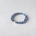 A beaded stretch bracelet featuring small blue and gray beads with a single white teardrop pearl accent, photographed on a light gray textured background.