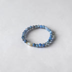A beaded stretch bracelet featuring small blue and gray beads with a single white teardrop pearl accent, photographed on a light gray textured background.