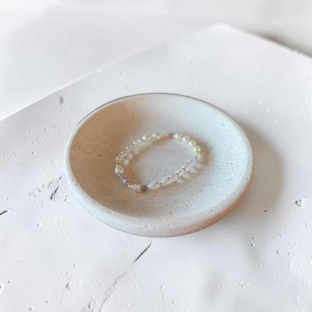 A delicate beaded bracelet with small clear and iridescent beads rests inside a shallow, round ceramic dish. The dish has a speckled off-white glaze with a warm beige rim, set against a textured white surface.