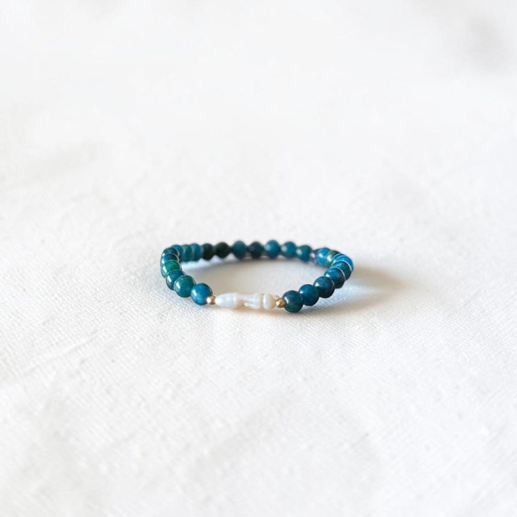 A beaded stretch bracelet featuring dark teal and turquoise beads with a small white pearl accent, resting on a textured white fabric background.