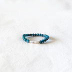 A beaded stretch bracelet featuring dark teal and turquoise beads with a small white pearl accent, resting on a textured white fabric background.