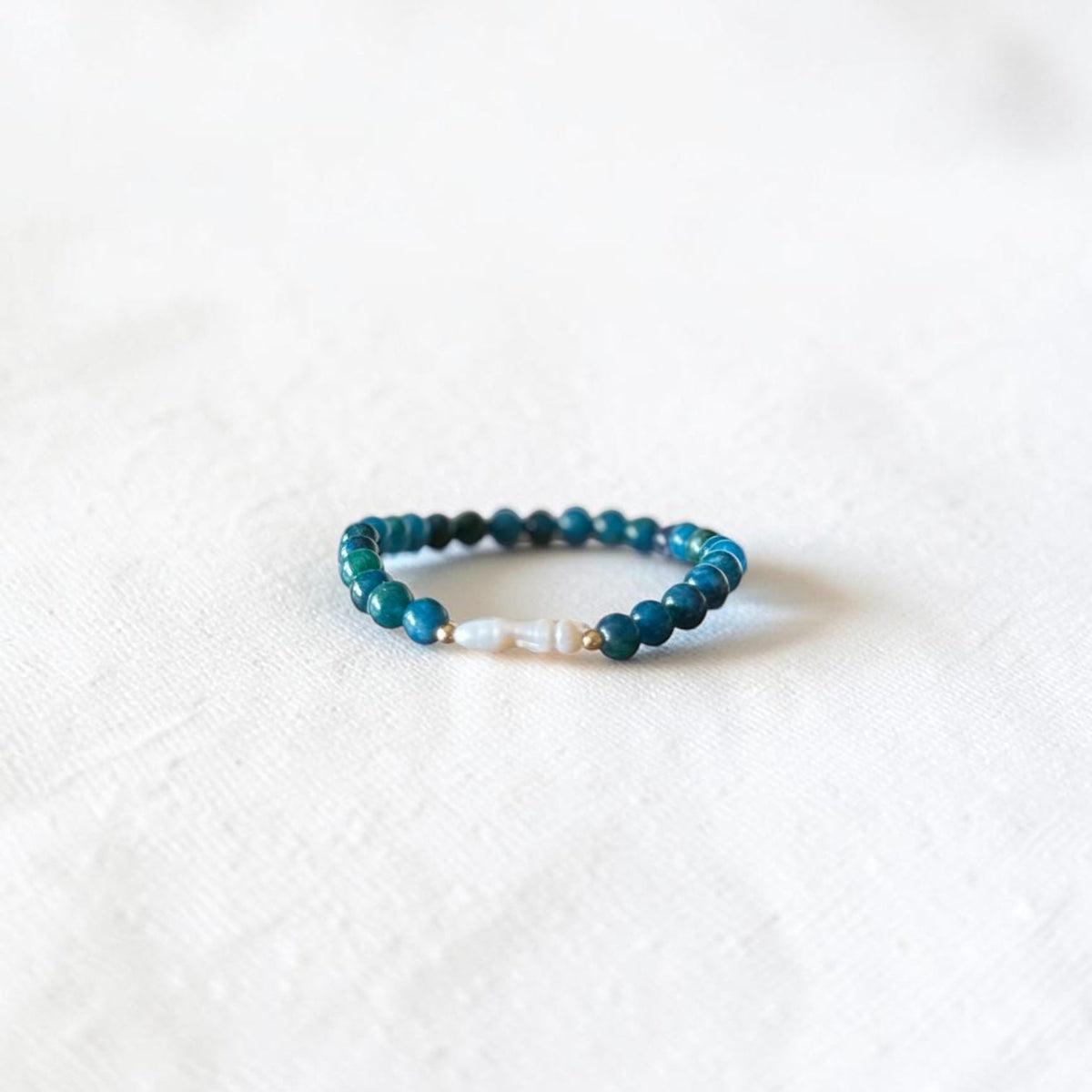 A beaded stretch bracelet featuring dark teal and turquoise beads with a small white pearl accent, resting on a textured white fabric background.